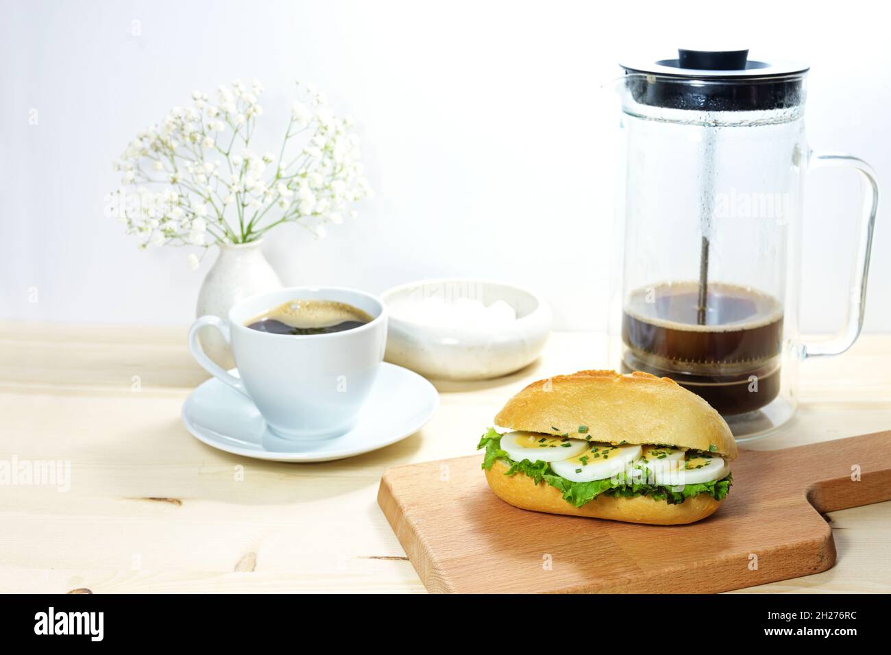 Colazione con fette di uova in un panino su un asse da cucina in legno, macchina per il caffè e tazza, sfondo chiaro con spazio copia, fuoco selezionato, stretto Foto Stock
