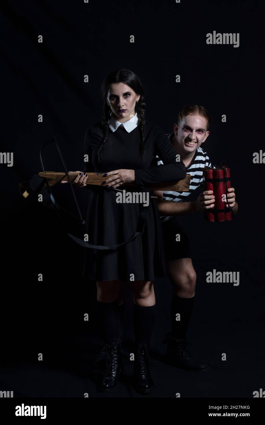 giovane donna scura e pallida vestita di nero con una balestra in mano fissando dritto davanti, dietro di lei un giovane sorridente con pochi sti Foto Stock