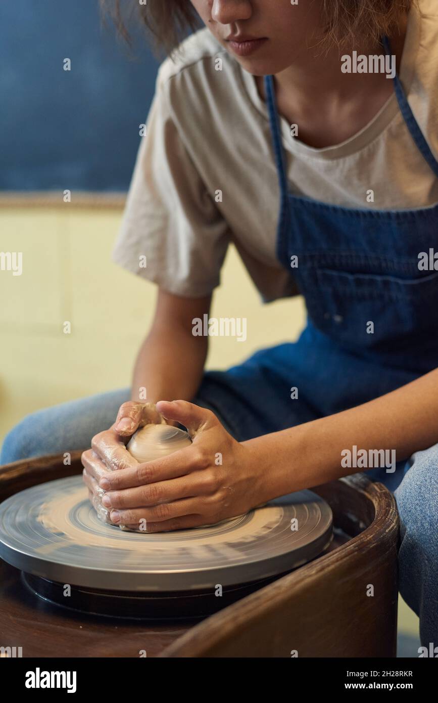 Primo piano di una ragazza seria in grembiule che tiene le mani sull'argilla mentre la forma sulla ruota che gira in officina Foto Stock