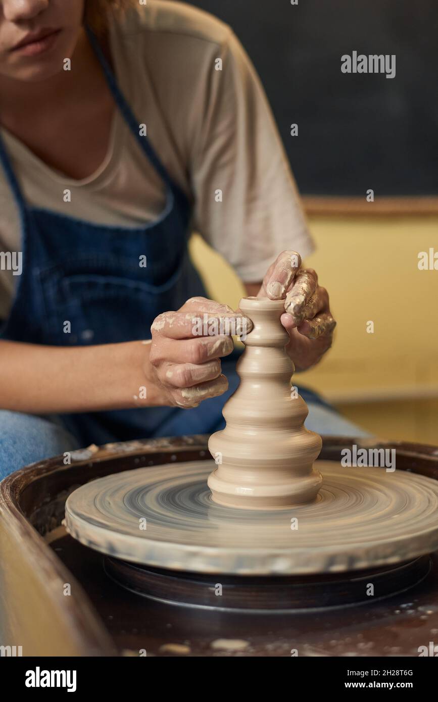 Primo piano di una ragazza irriconoscibile in grembiule che scolpisce il bel vaso di argilla con le mani sporche sulla ruota in ceramica Foto Stock