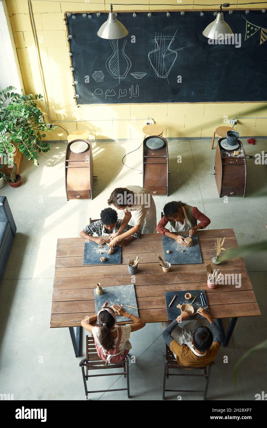 Direttamente sopra la vista di bambini multietnici che fanno vasi fatti a mano sotto il controllo dell'insegnante alla classe di ceramica Foto Stock