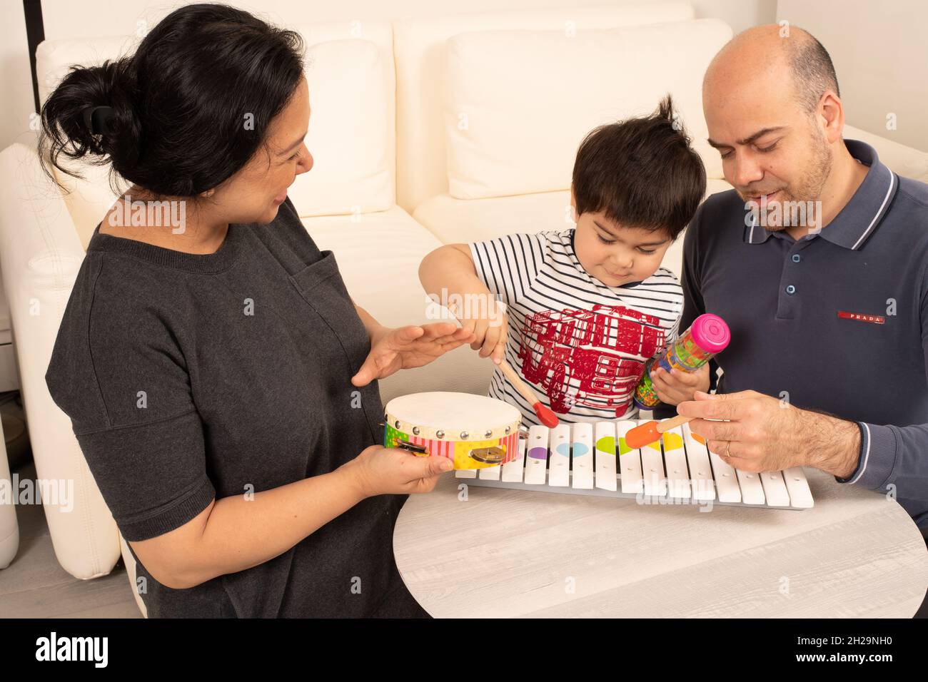 bambino ragazzo di 2 anni che suona lo xilofono di legno con un mazzuolo, madre e padre che fa musica con i hiim usando il tambourine e un altro mazzuolo Foto Stock