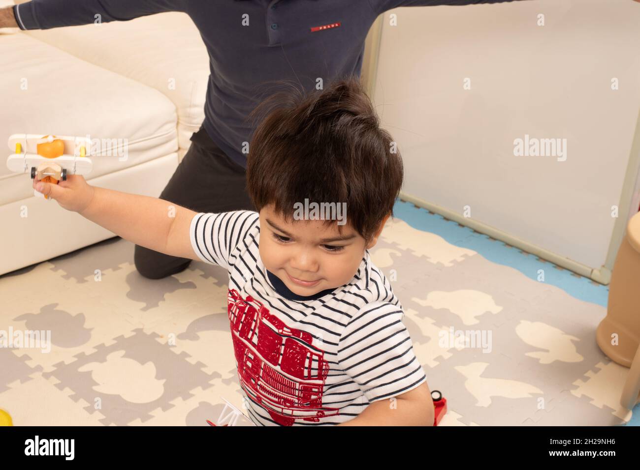 bambino di 2 anni a casa volare giocattoli aeroplano nelle sue mani, padre in background Foto Stock