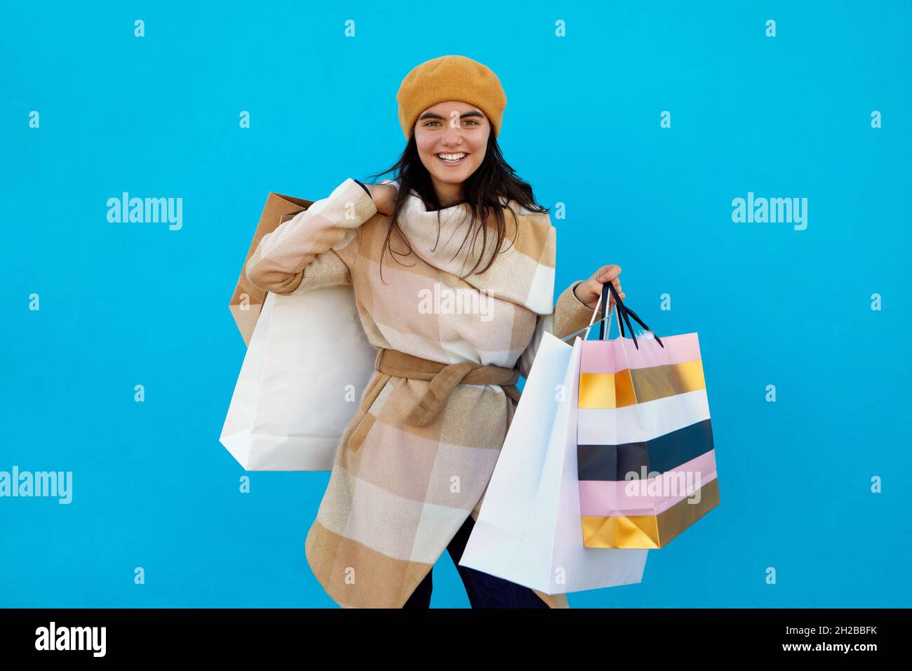 Cute attraente felice sorridente elegante donna shopaholic in elegante cappotto in plaid beige e beretto che tiene borse shopping su sfondo blu studio isolato, Foto Stock