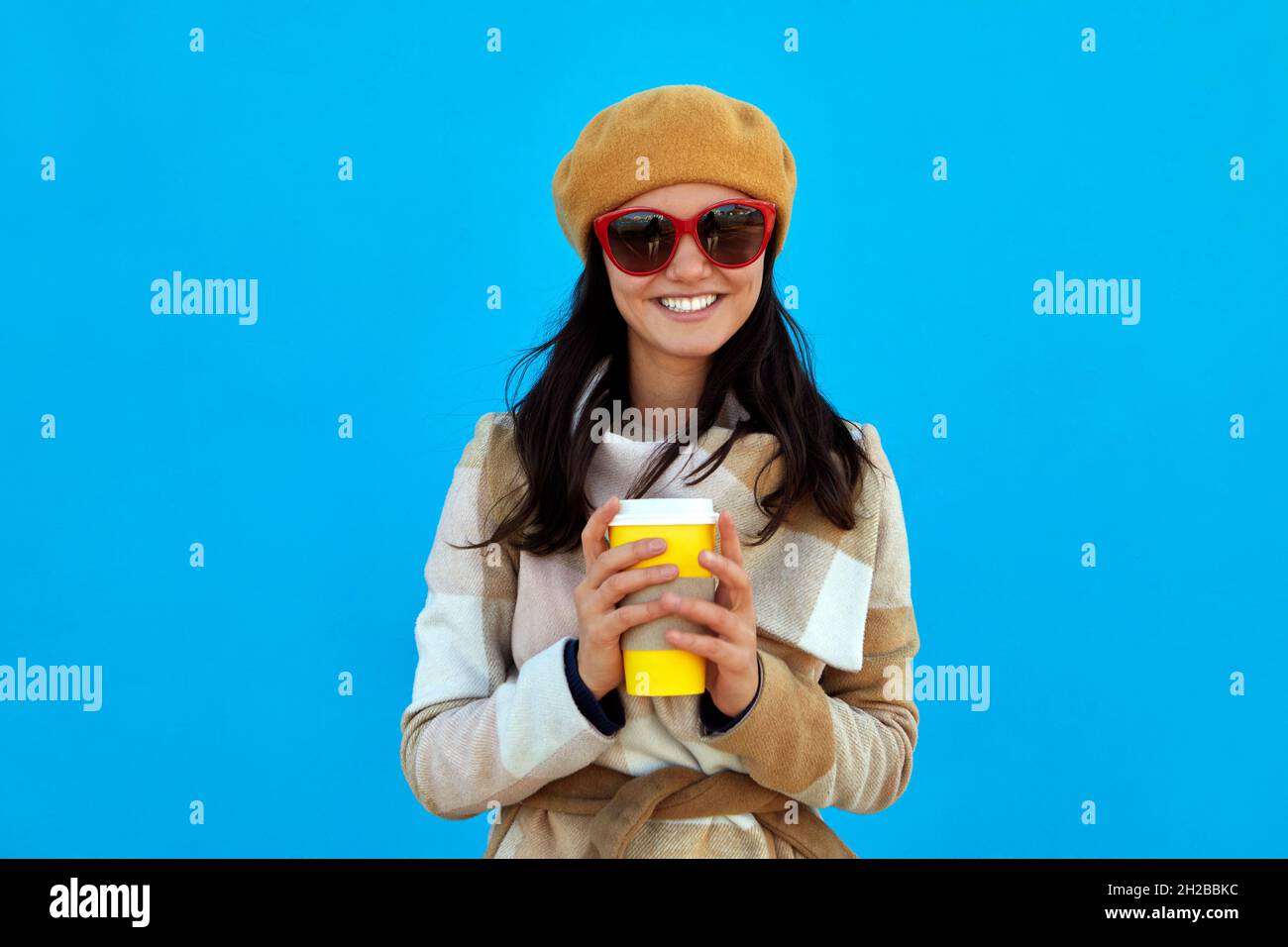 Emozionale felice bella donna in beretta beige cappotto e occhiali da sole su sfondo blu bere caffè in carta tazza sorridente, cibo del mattino, bevanda gioiosa Foto Stock