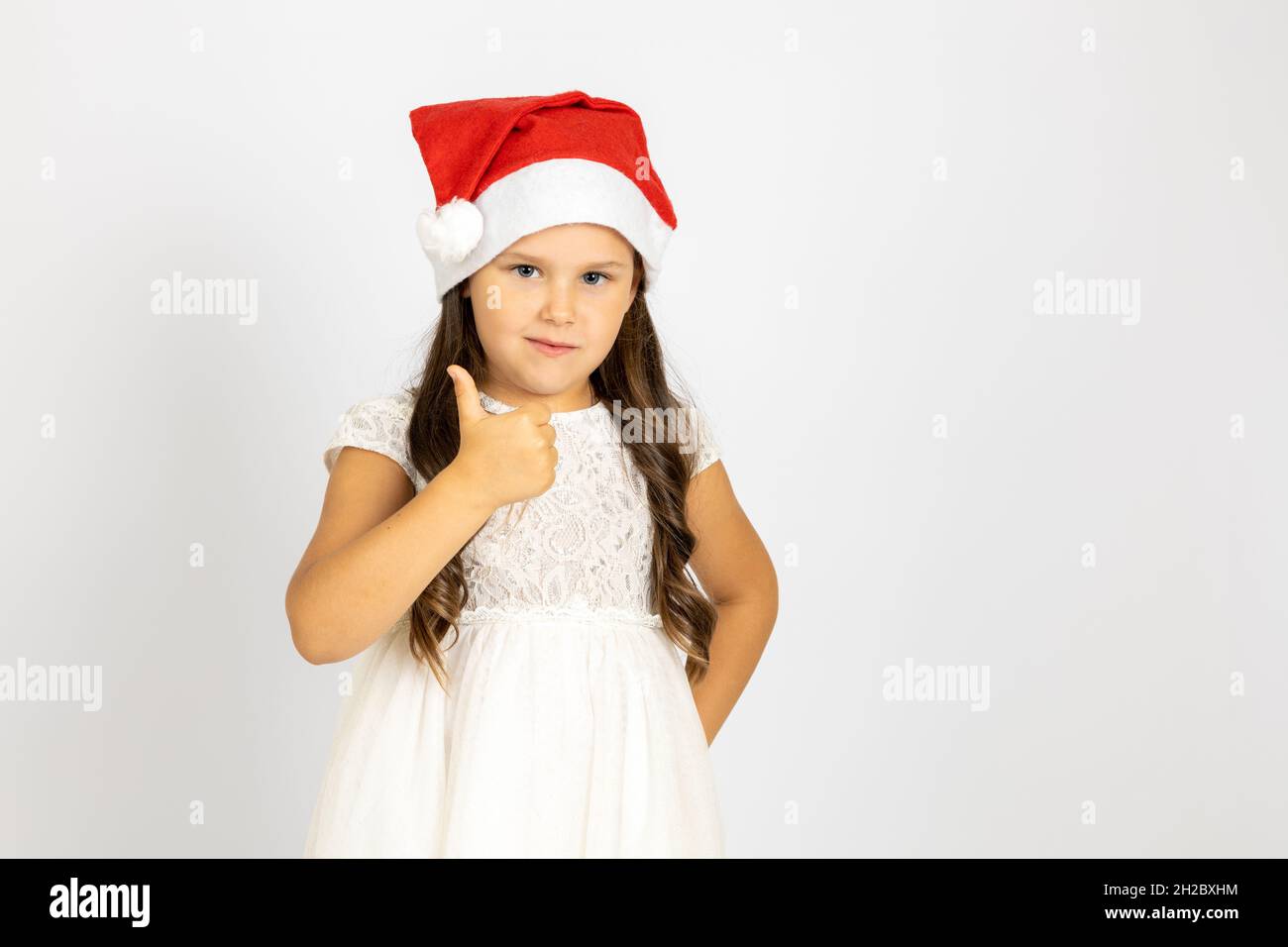 Ritratto di sorridente, entusiasta bambino in rosso Babbo Natale cappello dà i pollici in su, isolato su sfondo bianco con spazio copia Foto Stock