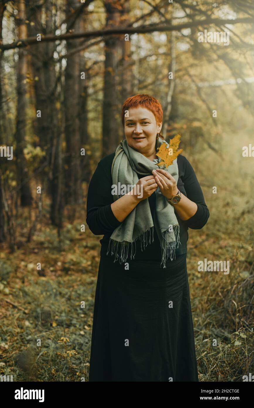 Sorridente donna di mezza età 30-40 anni cammina nella foresta autunnale con una foglia di acero giallo in mano Foto Stock