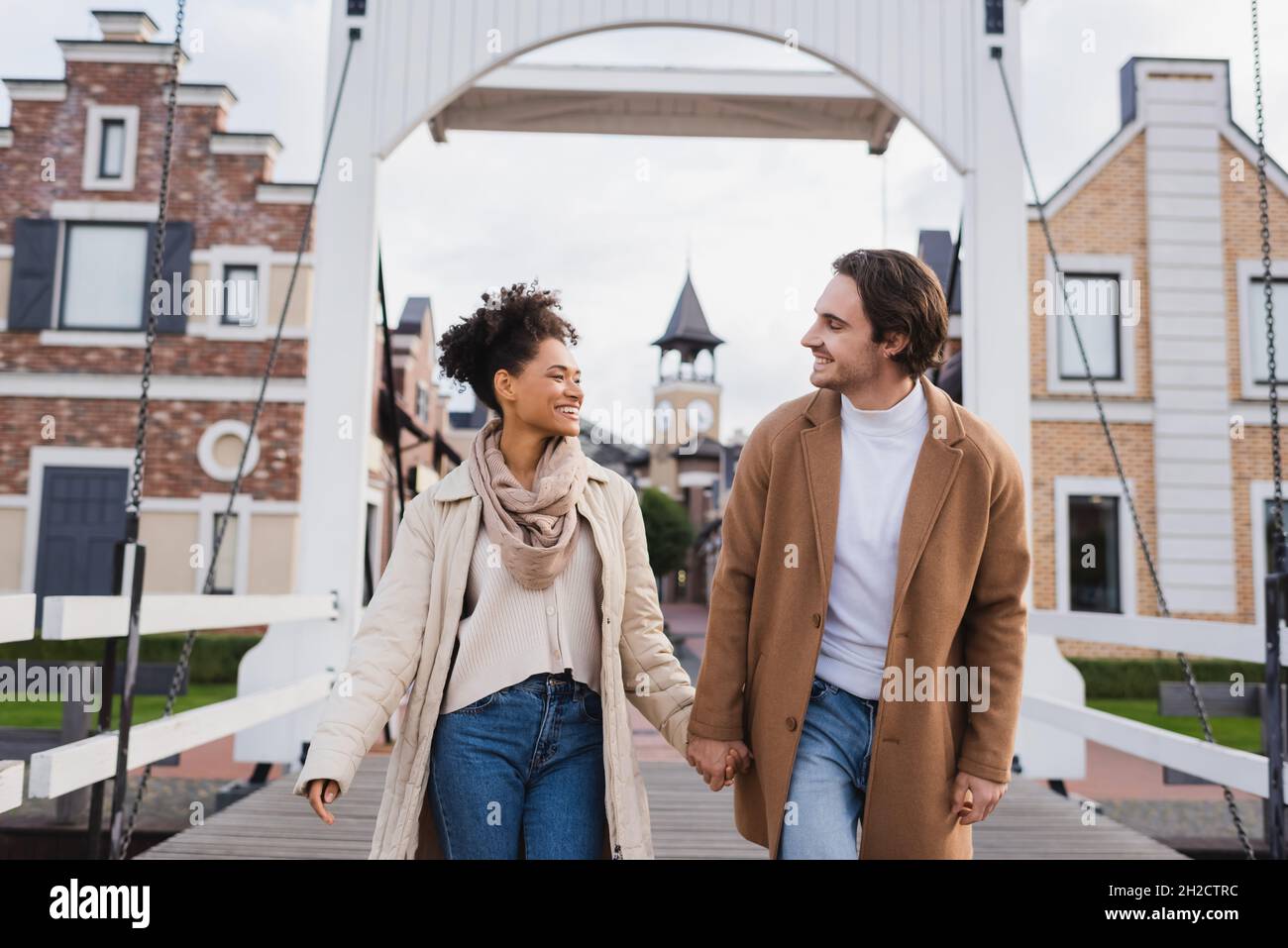 sorridente coppia multietnica in cappotti che tengono le mani vicino al centro commerciale Foto Stock