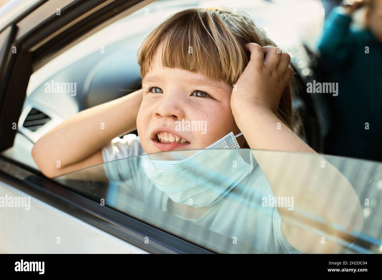 Il bambino si siede in auto tenendo una maschera e guarda lontano dalla finestra Foto Stock