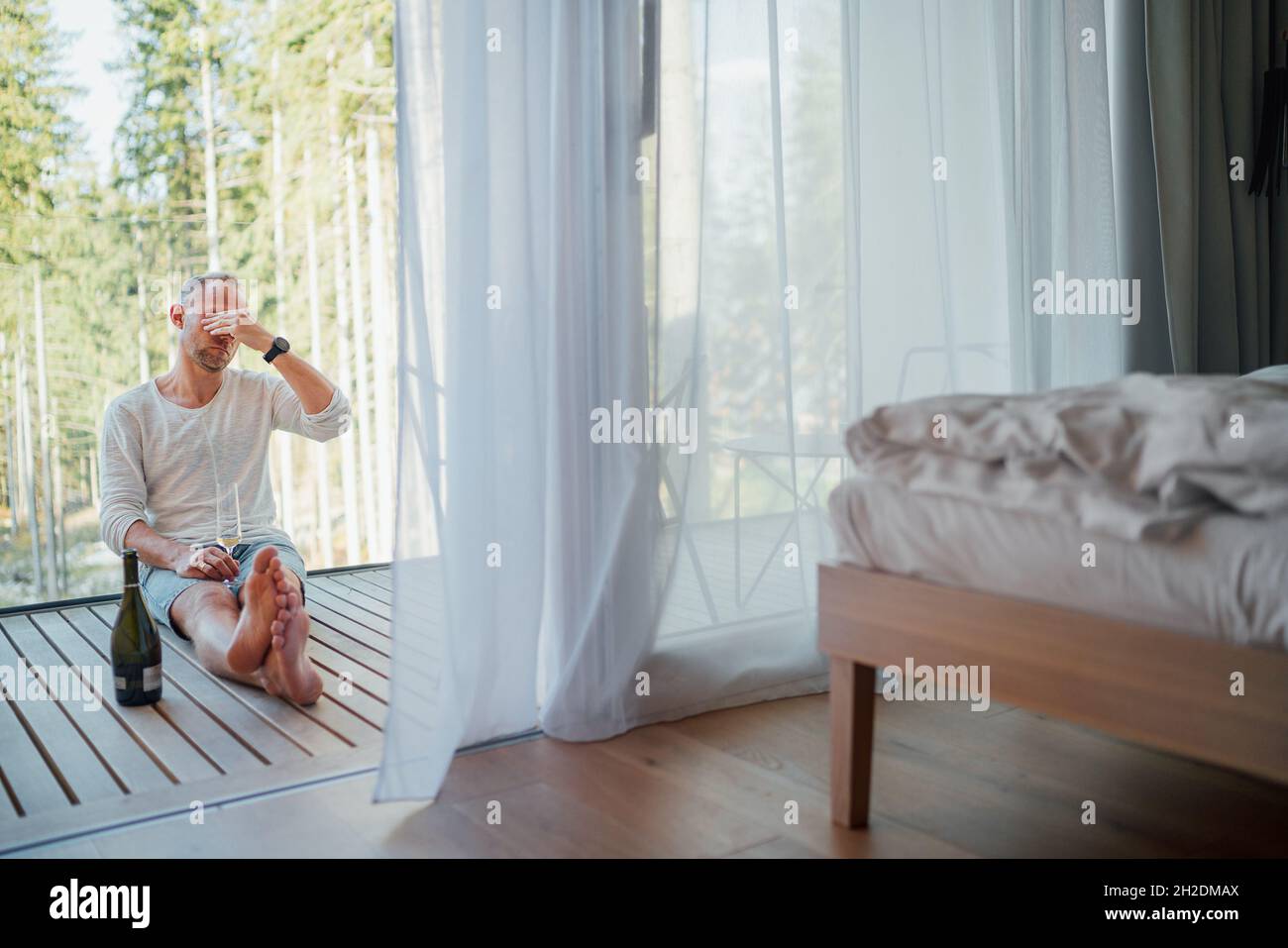 Ritratto di un uomo stanco di mezza età seduto a piedi nudi sul pavimento della casa forestale balcone, egli bere vino bianco e massaggiando templi. Persone esauste o m Foto Stock