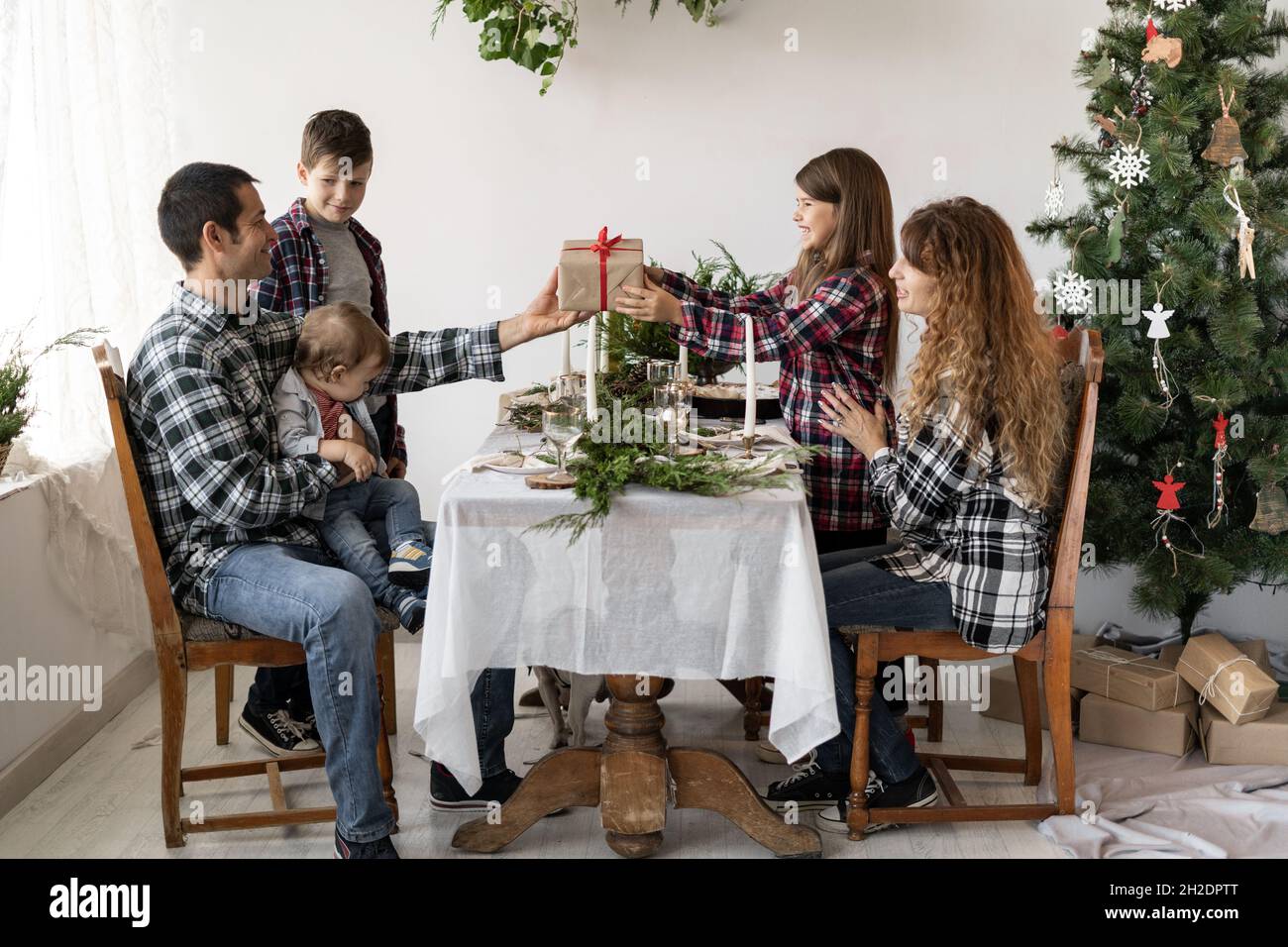 La grande famiglia ha una colazione di Natale al tavolo nel soggiorno e papà dà una scatola con un regalo a una bambina per il nuovo anno. Tavolo delle feste Foto Stock