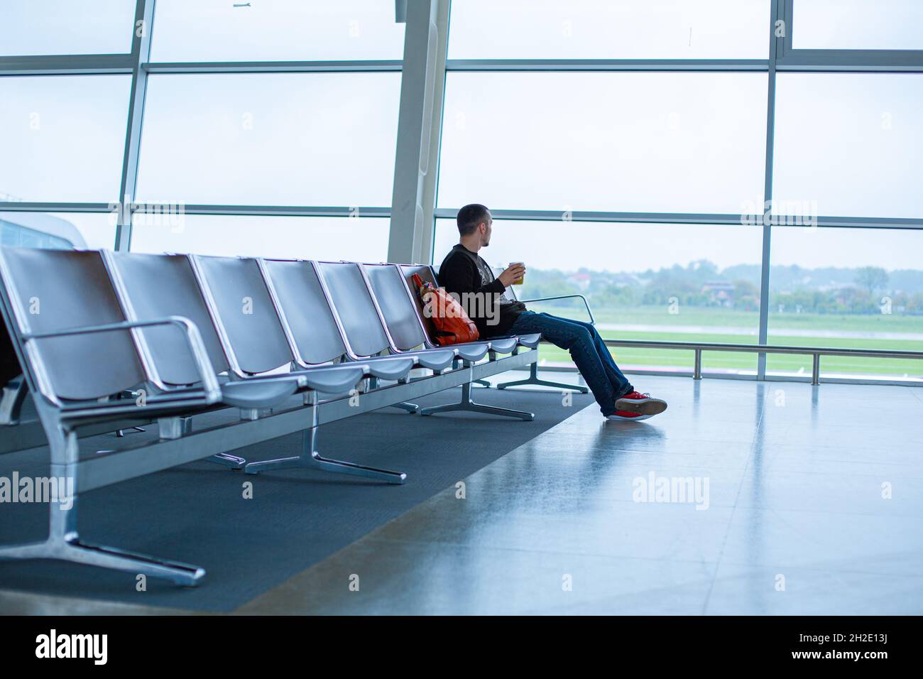 Un ragazzo è seduto su una fila vuota di posti a sedere di fronte a una  grande finestra di vetro colorato in un terminal dell'aeroporto, in attesa di  un volo Foto stock -