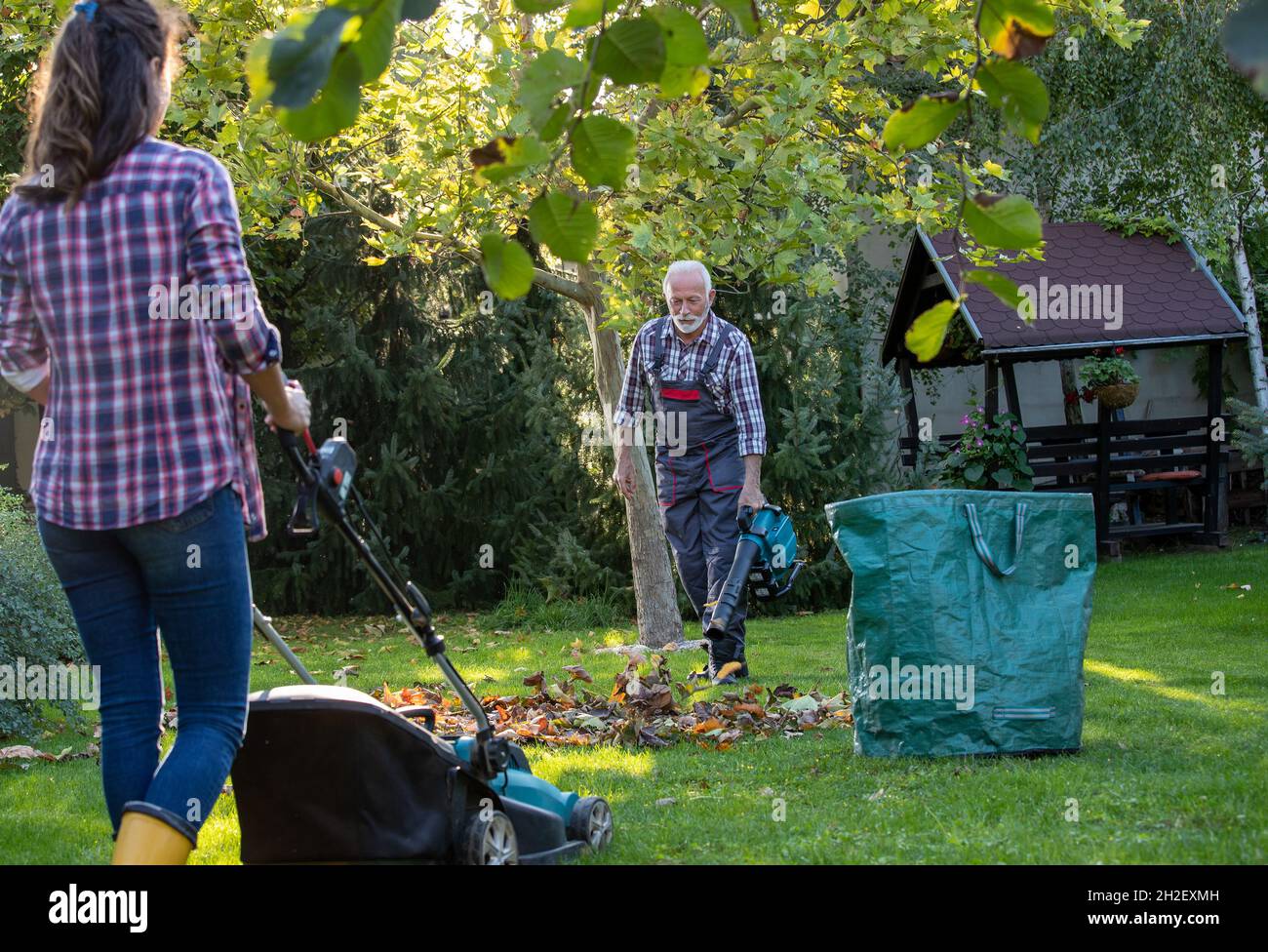 Uomo anziano nel lavoro generale in giardino con soffiatore di foglie mentre la figlia falciando prato in autunno Foto Stock