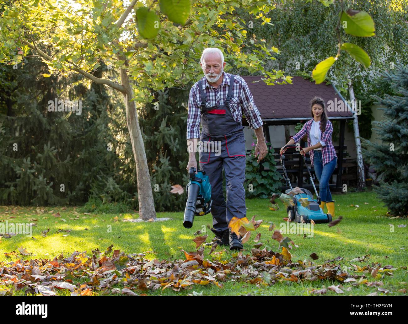 Uomo anziano in generale lavorando in giardino con soffiatore foglia e figlia falciando prato in background in autunno Foto Stock