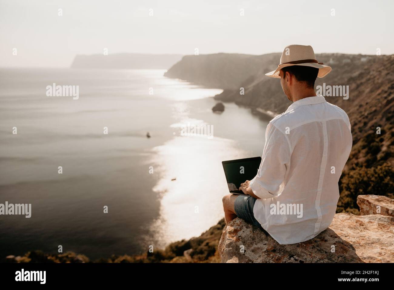 Digital nomad, uomo nel cappello, un uomo d'affari con un laptop siede sulle rocce vicino al mare durante il tramonto, effettua una transazione di affari in linea da a. Foto Stock