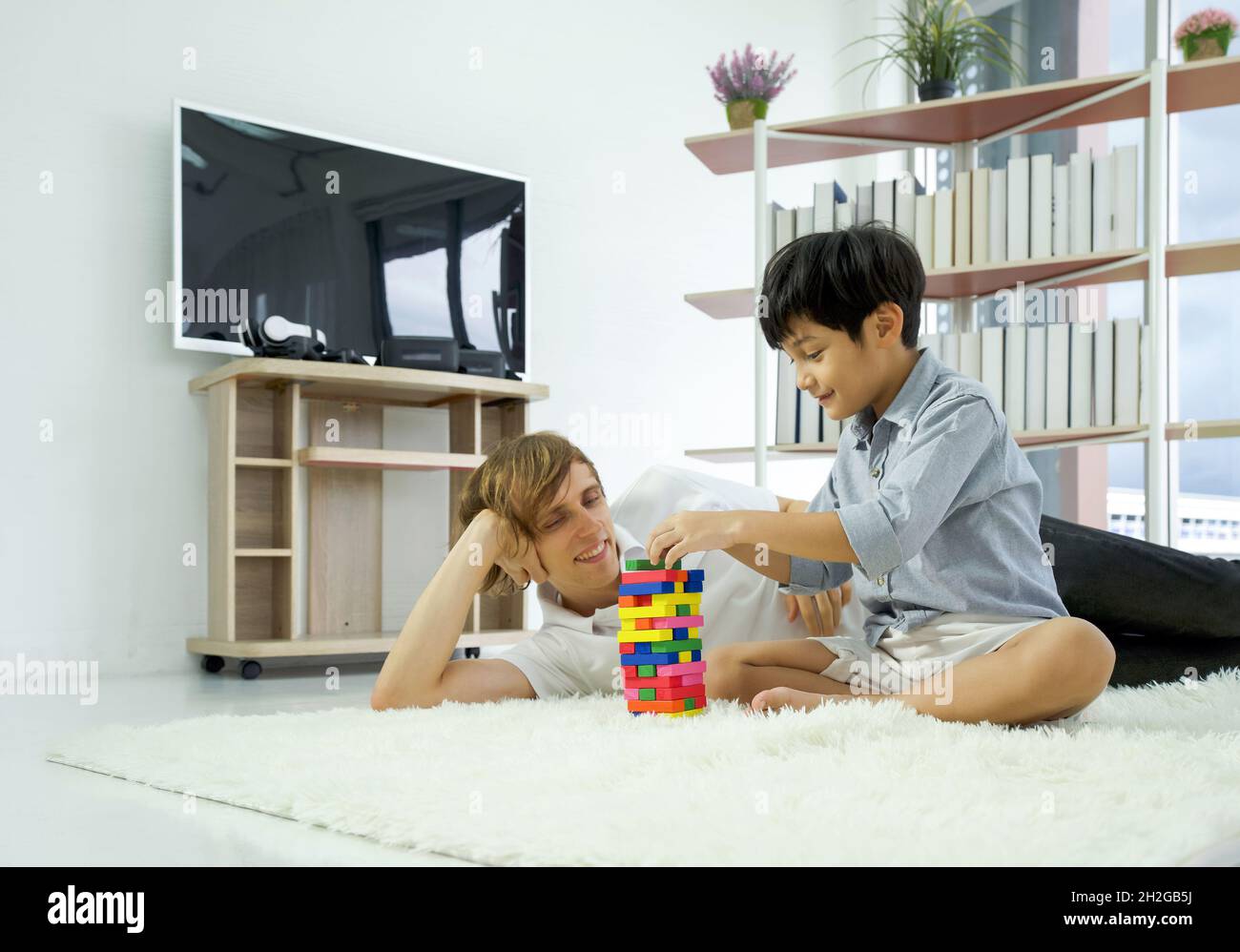 Il giovane padre caucasico e il suo figlio adottivo godono di giocare i blocchi di legno giocattolo nel soggiorno durante le vacanze. Equilibrio della vita lavorativa. Foto Stock