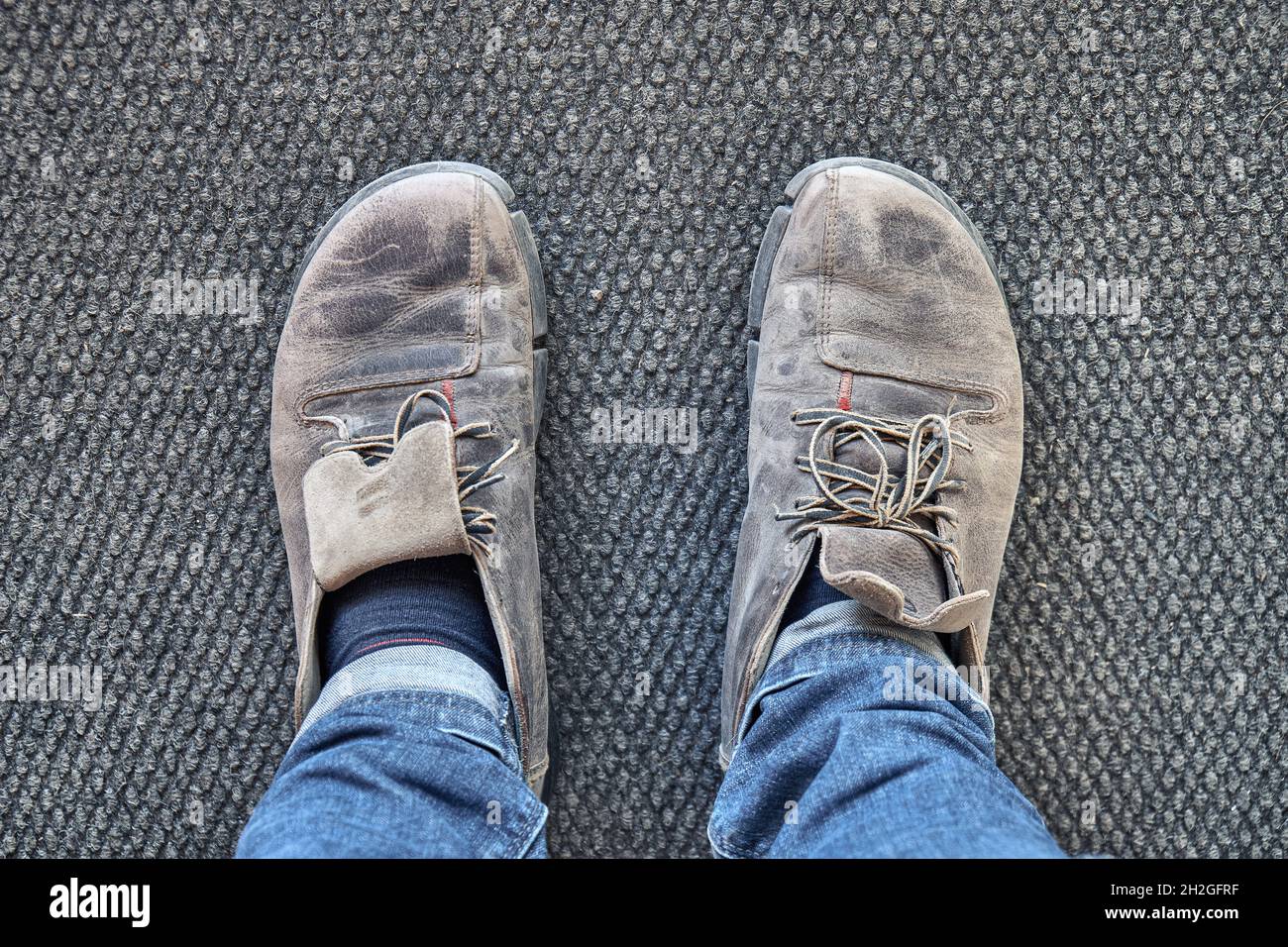 Le gambe dell'uomo in jeans blu e vecchie scarpe marroni si erigono su tappeto grigio nella vista del primo punto dell'officina Foto Stock