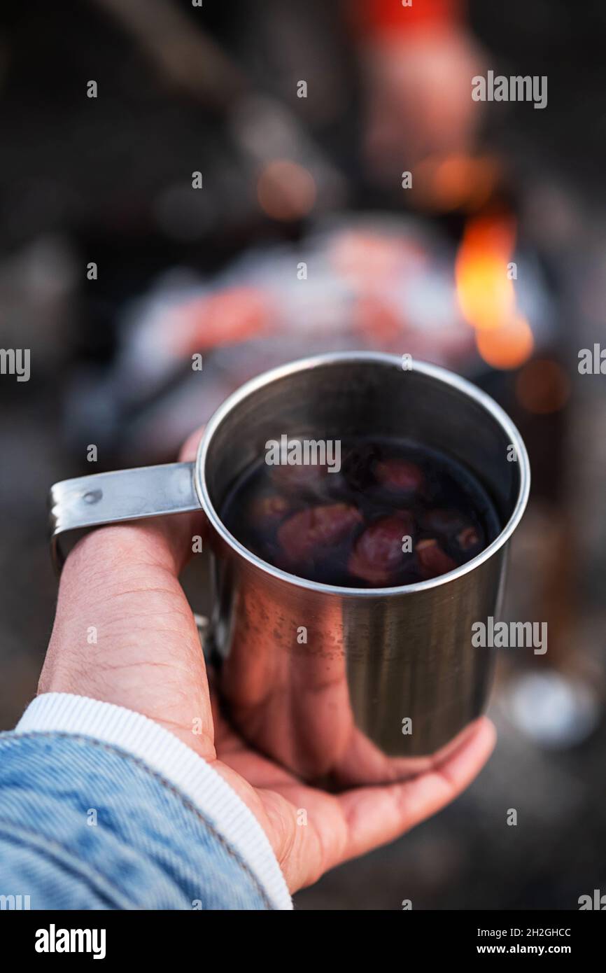 Donna mano tenere rosa hip tè in tazza di metallo contro falò. Turismo ecologico, stile di vita sostenibile. Foto Stock