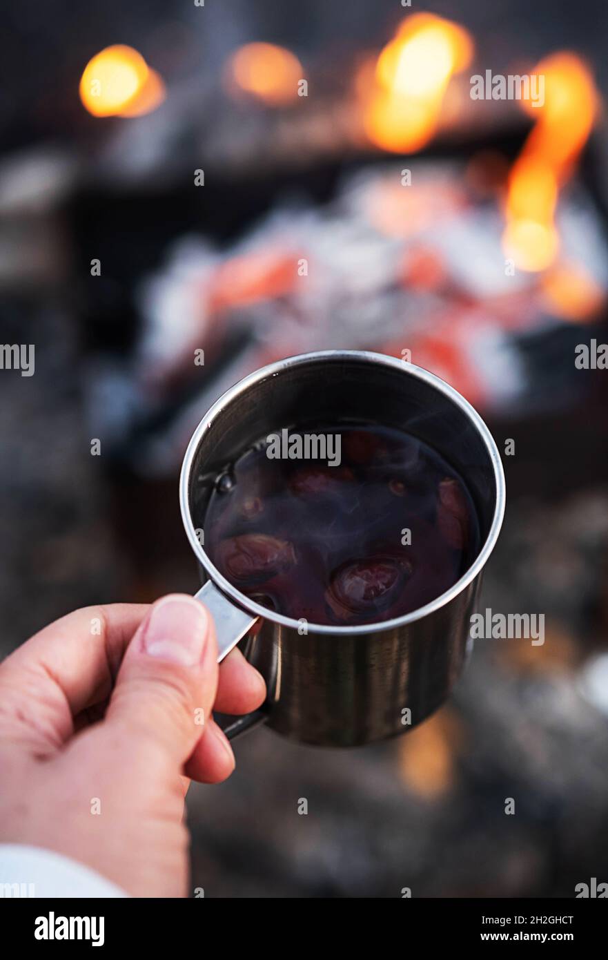 Donna mano tenere rosa hip tè in tazza di metallo contro falò. Turismo ecologico, stile di vita sostenibile. Foto Stock