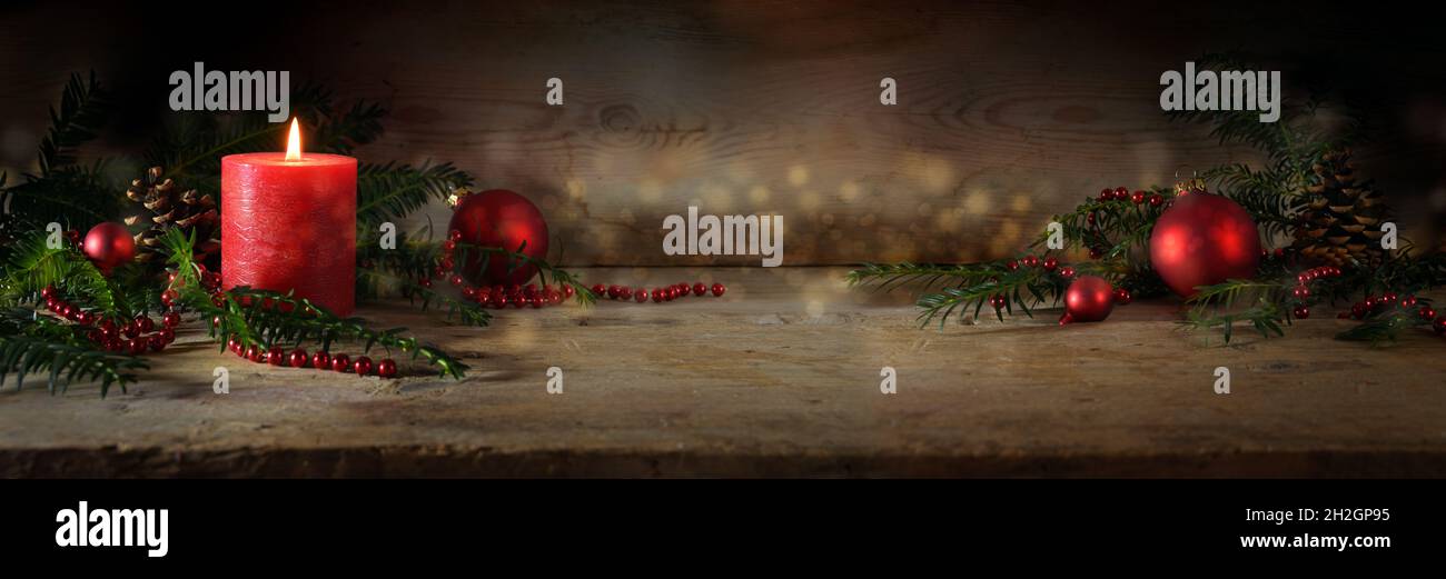 Decorazioni suggestive di Avvento e Natale con una candela rossa illuminata, palle e rami di tasso sempreverdi su tavole di legno rustico scuro, ampia forma panoramica Foto Stock