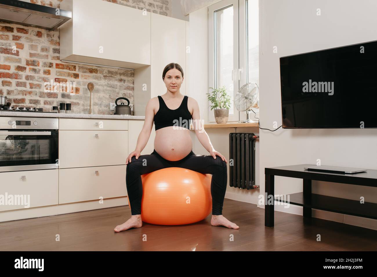 Una donna incinta è seduta su una palla arancione esercizio e rilassarsi dopo un allenamento fitness vicino alla cucina a casa sua. Gravidanza sana. Foto Stock