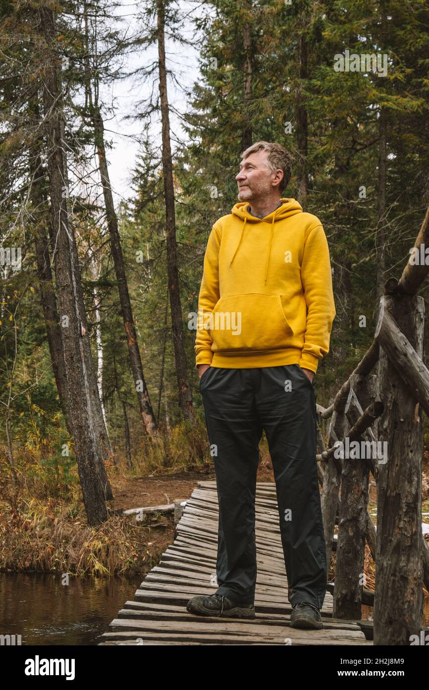 Un viaggiatore di mezza età in felpa con cappuccio gialla si trova in piedi sul ponte sopra un fiume nella foresta, godendo la splendida giornata autunnale all'aperto. Viaggi locali. Foto Stock