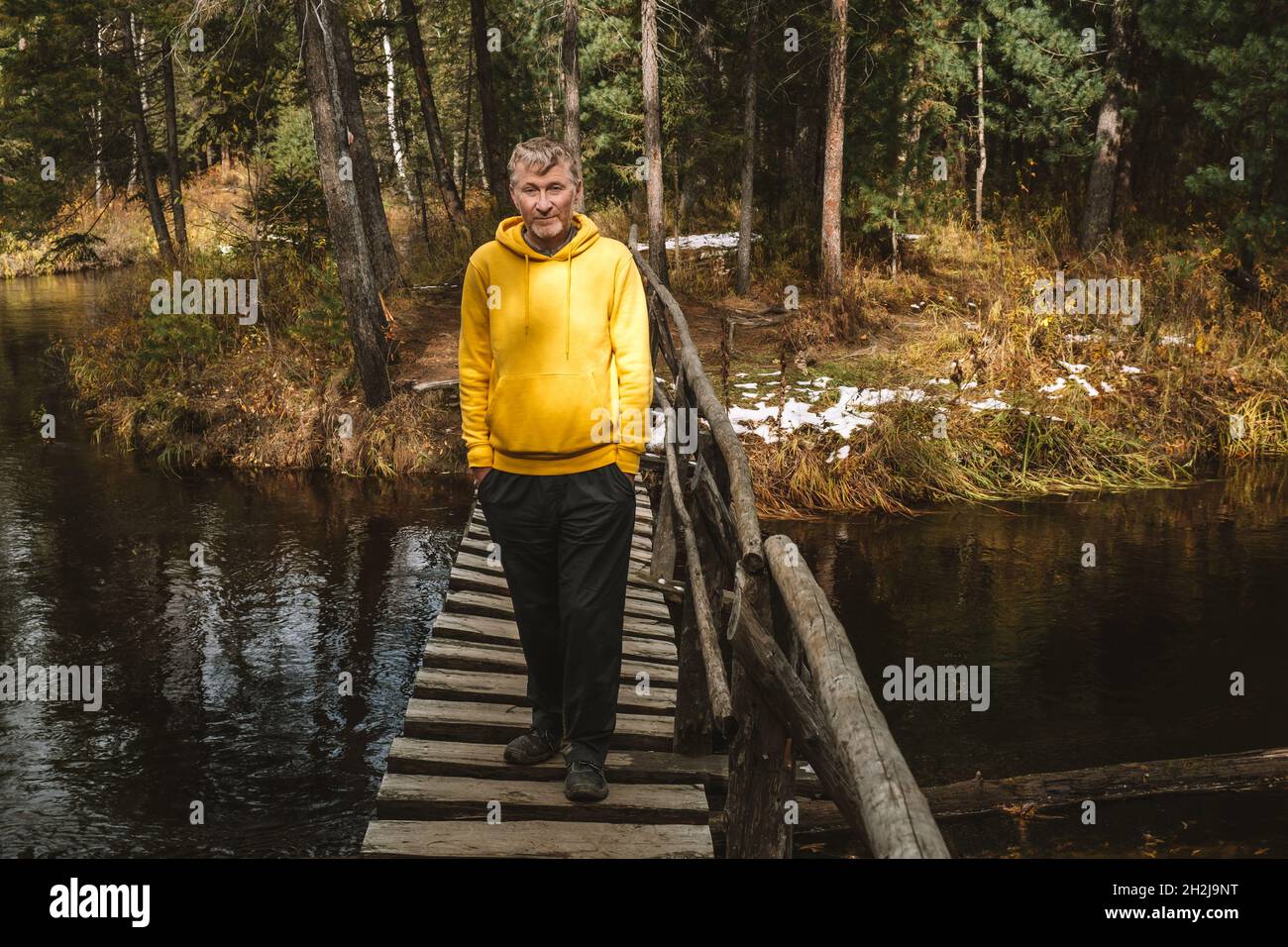 Un viaggiatore di mezza età in una felpa con cappuccio gialla attraversa un ponte di legno su un fiume in una foresta autunnale. Escursioni locali e passeggiate nel fine settimana. Foto Stock