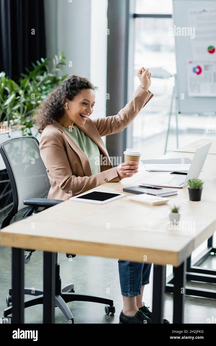 donna d'affari afroamericana con bevanda di takeaway che mostra il gesto di trionfo vicino al laptop in ufficio Foto Stock