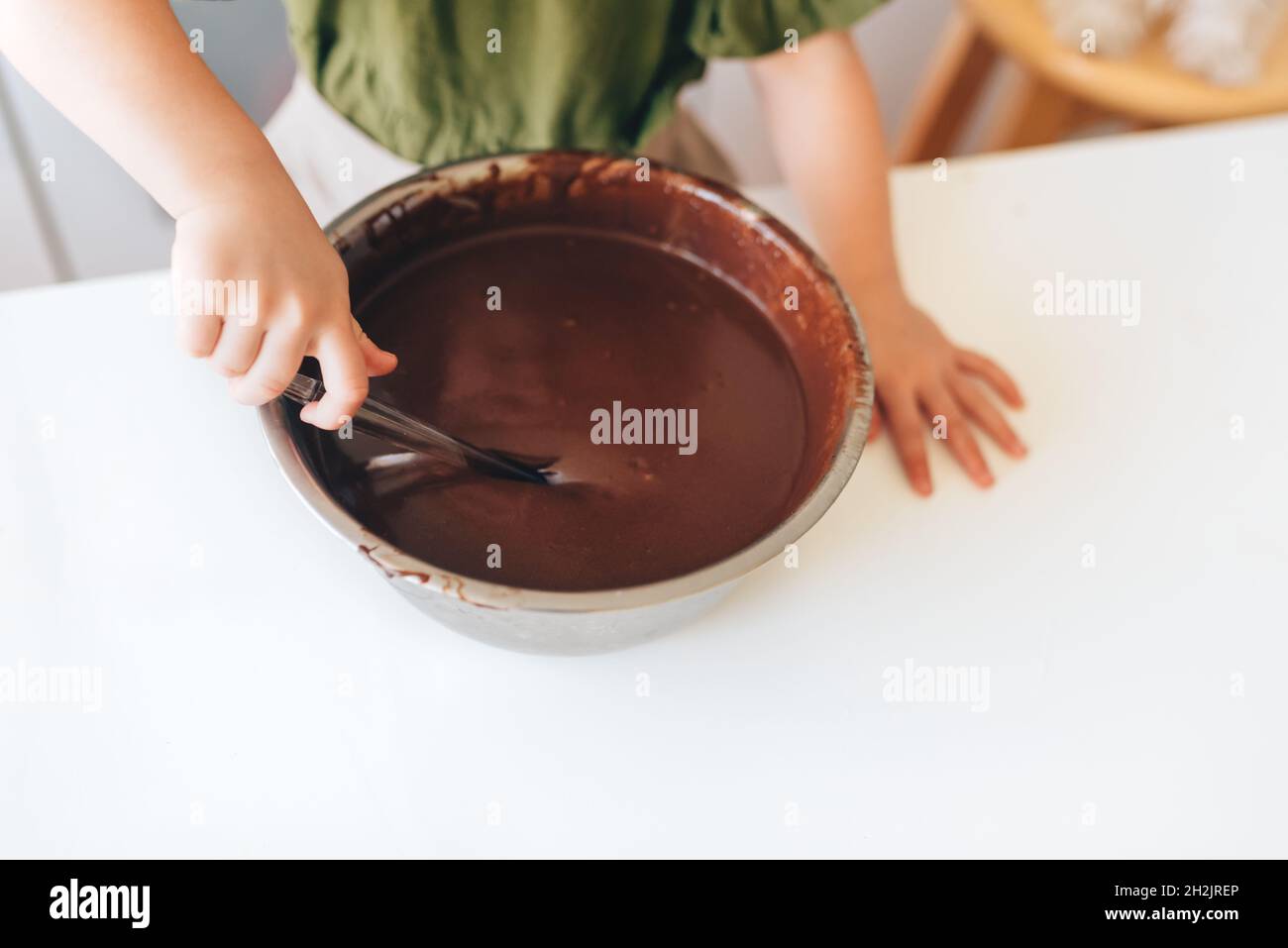 Il bambino che fa la pasta tradizionale al cioccolato per i biscotti di Natale su un tavolo bianco in cucina. Recipiente in metallo e cucchiaio in silicone trasparente. Foto Stock