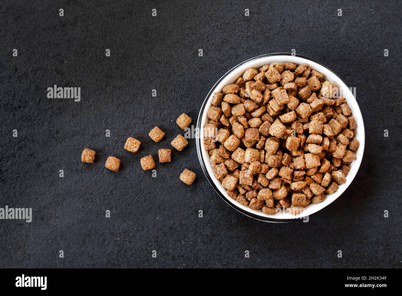 Asciugare gli alimenti per animali domestici in un recipiente in ceramica bianca su sfondo nero. Disposizione piatta, vista dall'alto, spazio di copia Foto Stock
