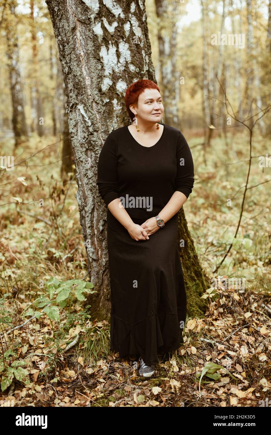 Una donna di mezza età di 30-40 anni in una foresta autunnale si erge su un albero, pensivo, sognante. Foto a tutta lunghezza Foto Stock