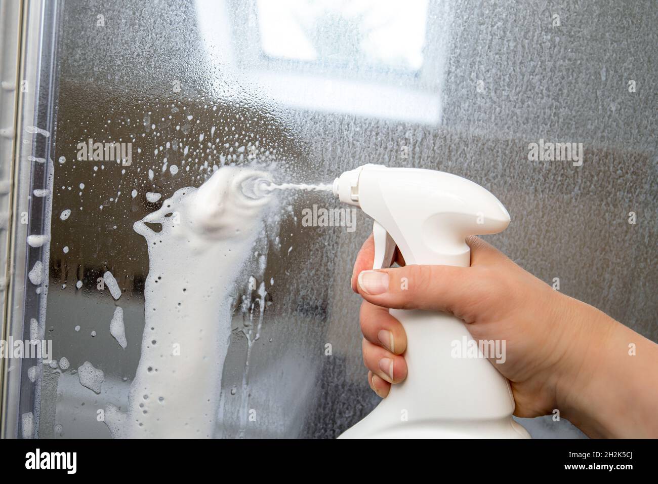 La mano della persona utilizza schiuma chimica per rimuovere le calcare. Rimozione del calcare dalla porta in vetro del box doccia. Lavori in corso. Foto Stock