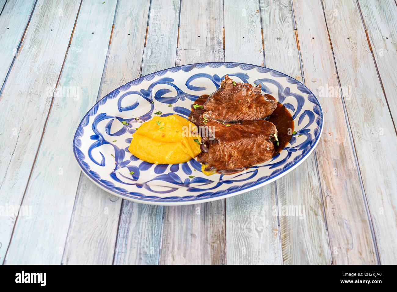 Deliziosa ricetta spagnola per guanciale di manzo stufato in salsa e purea di patate dolci con prezzemolo su un piatto di porcellana blu di Talavera Foto Stock