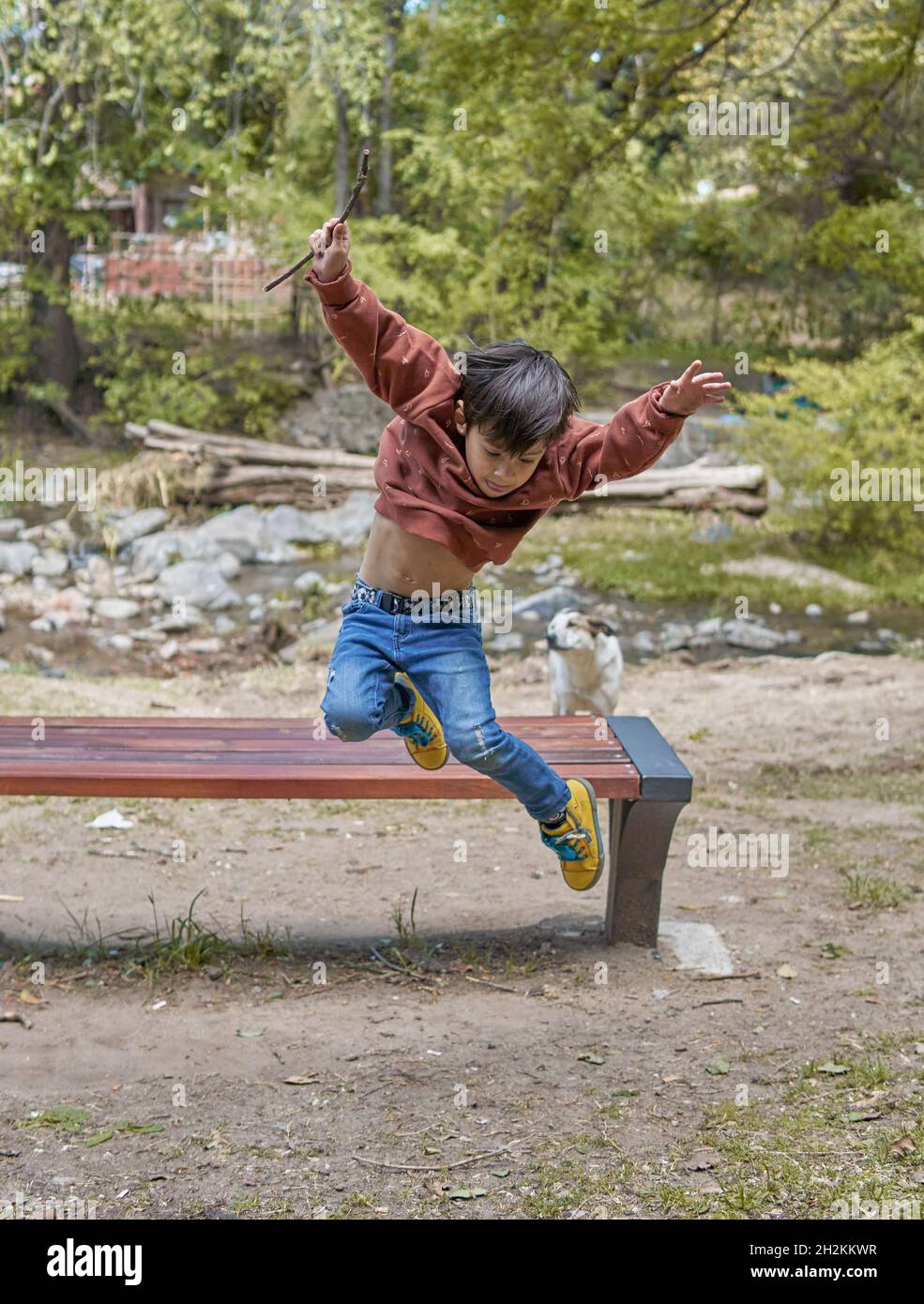 bambino latino che salta da una panca di legno a lato del torrente con le braccia e le gambe sollevate nell'aria. Orizzontale. Argentina Foto Stock
