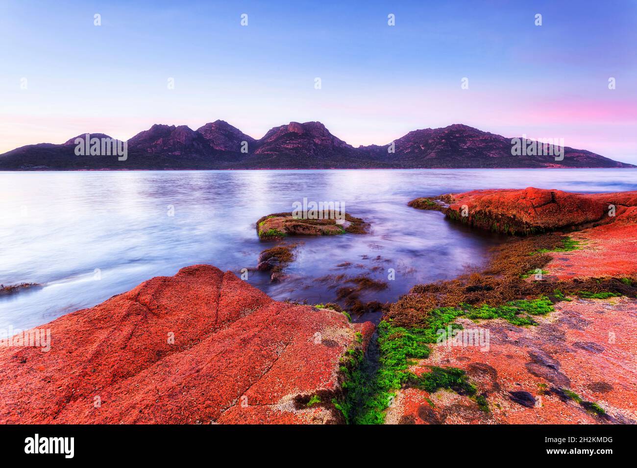 Rosa rosso licheni coperto rocce di masso di granito sulle rive della baia di Coles in Tasmania - alba panoramica. Foto Stock