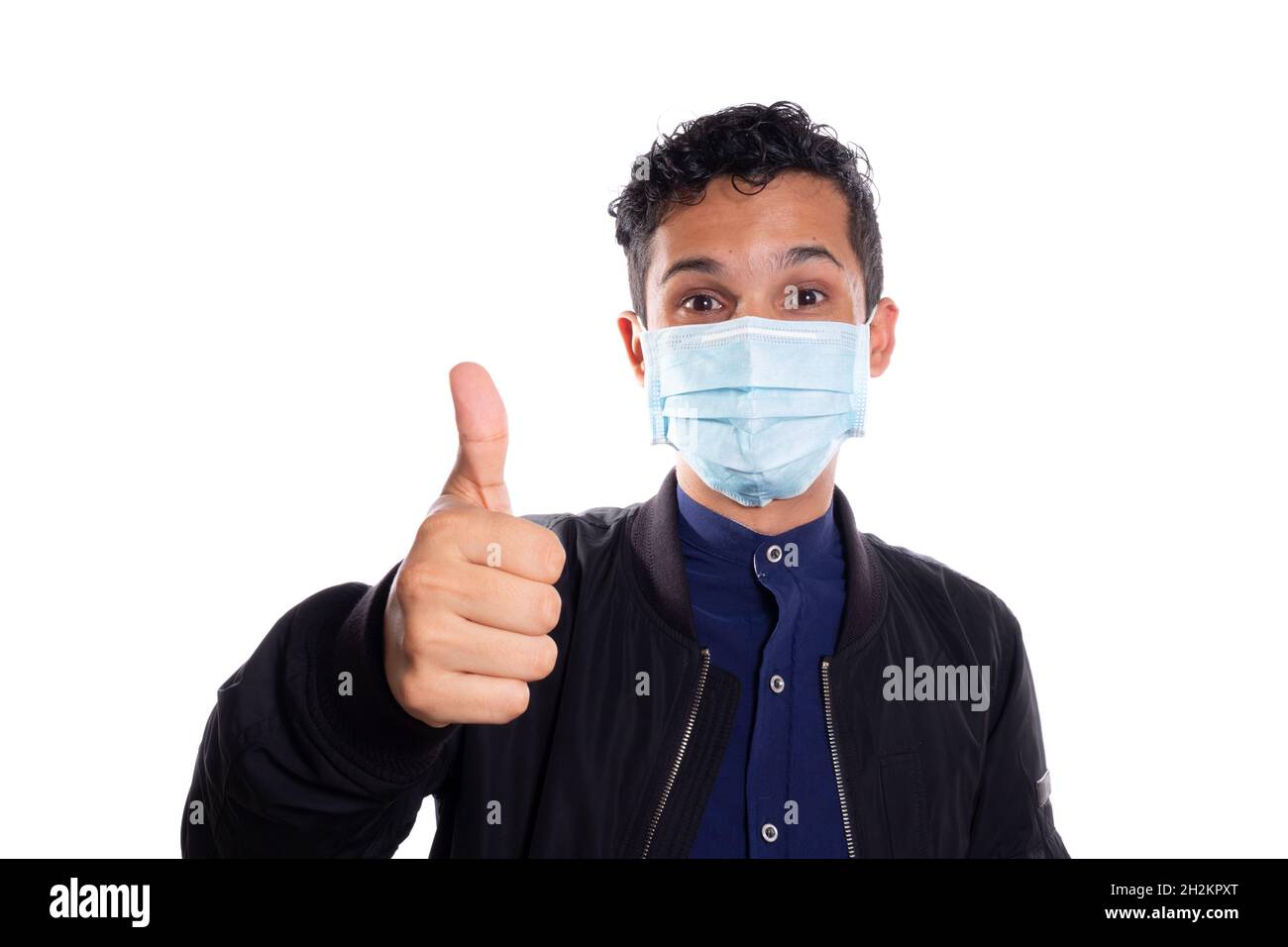 Uomo che indossa la maschera del viso tenendo il pollice della sua mano. Sfondo bianco. Giovane adulto latino giovane uomo che mostra i pollici in su. Foto Stock