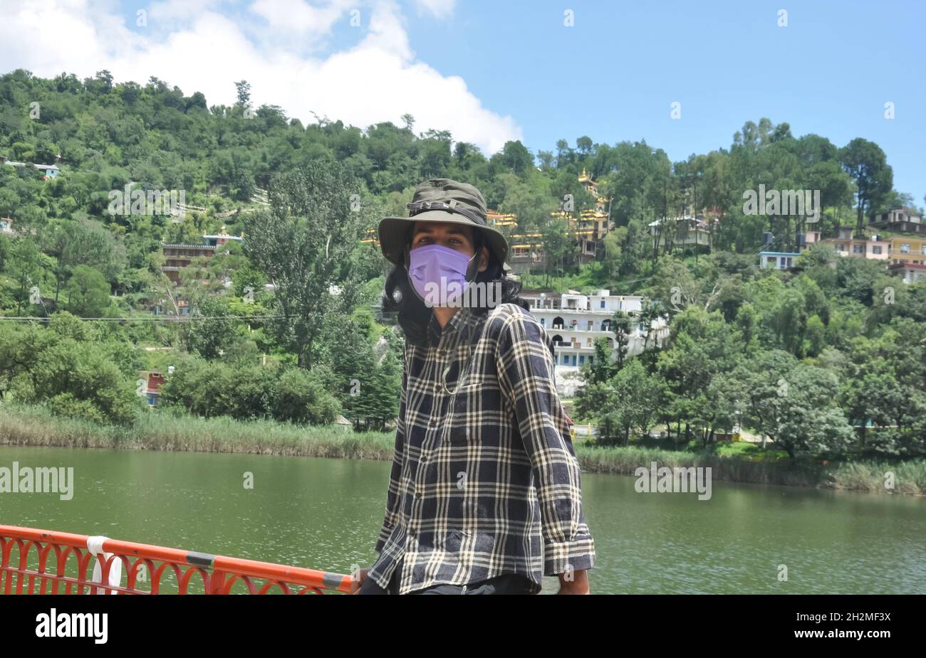 Un giovane uomo dai capelli lunghi che indossa cappello e maschera per il viso e guarda la macchina fotografica con seduta sulla barriera di sicurezza vicino al lago di Rewalsar (Tso-Pema) Foto Stock