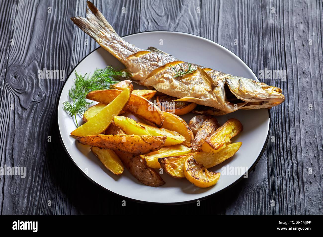 Pesce branzino arrosto servito su un piatto con sale marino, aneto, patatine fritte rafano su un tavolo di legno nero, vista dall'alto, primo piano Foto Stock