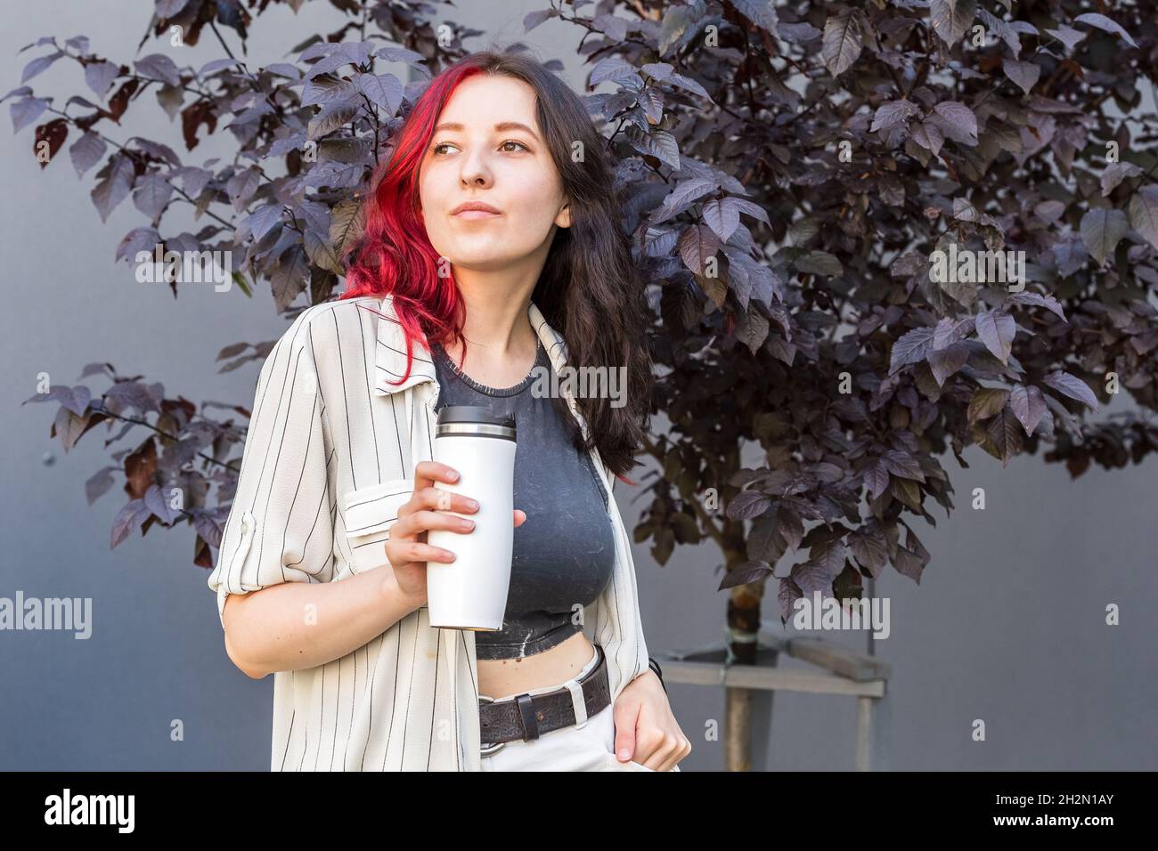 Giovane donna con capelli neri e rossi creativi e colorati che tiene una tazza bianca riutilizzabile isolata per il caffè per andare nelle sue mani. Foto Stock