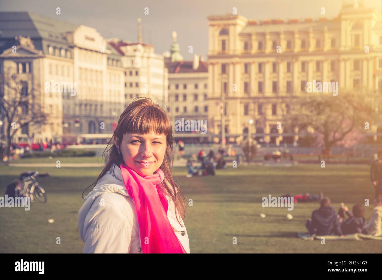 Giovane ragazza viaggiatore con giacca bianca guardando via e sorridere nel Parco Sigmund Freud del centro storico di Vienna con vecchi edifici sfondo, wom Foto Stock