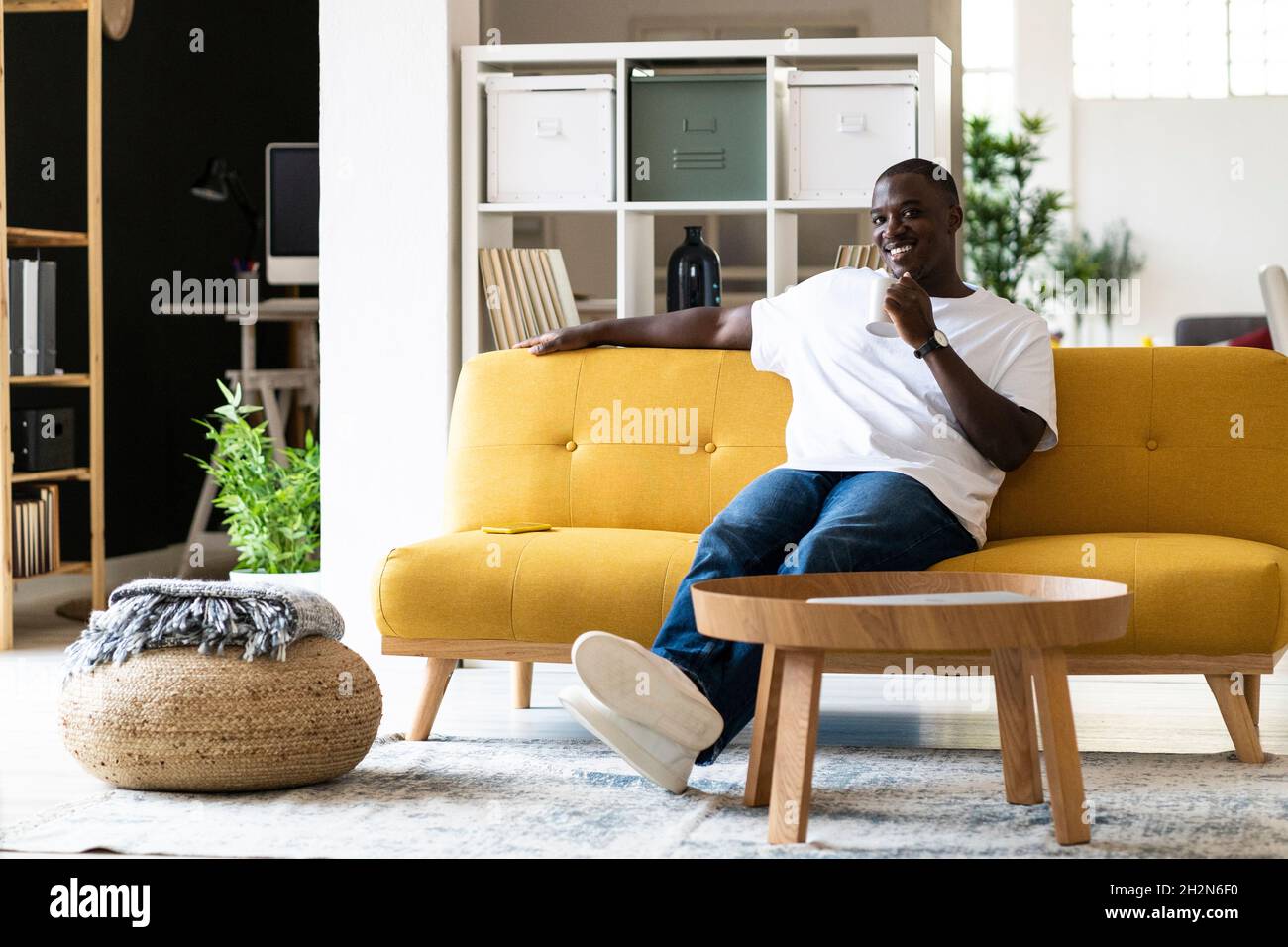 Uomo sorridente che tiene messaggi di testo con la tazza tramite smartphone mentre si siede sul divano nel soggiorno di casa Foto Stock