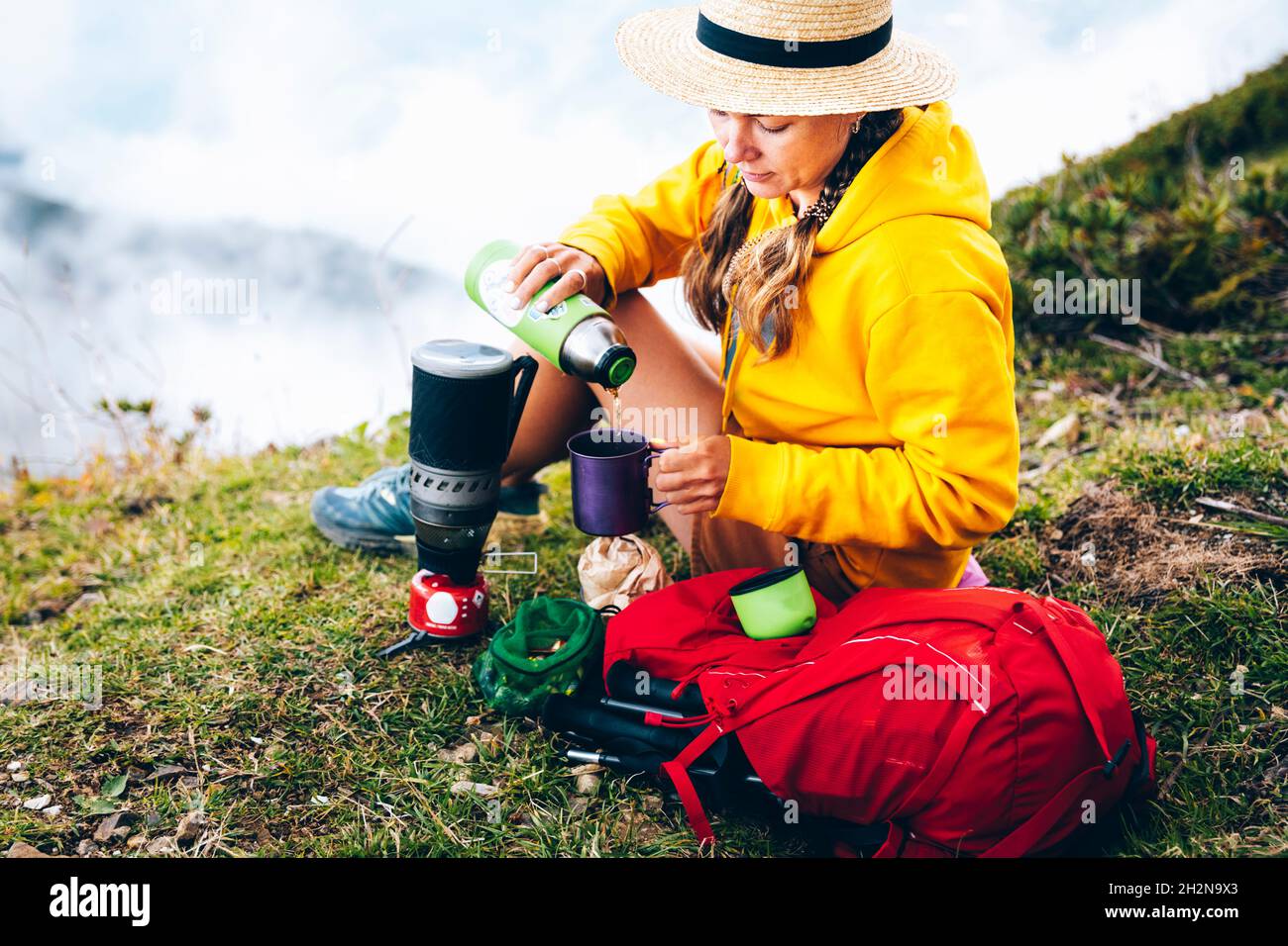 Donna che indossa un cappello che versa il tè in tazza mentre si siede da zaino in montagna Foto Stock