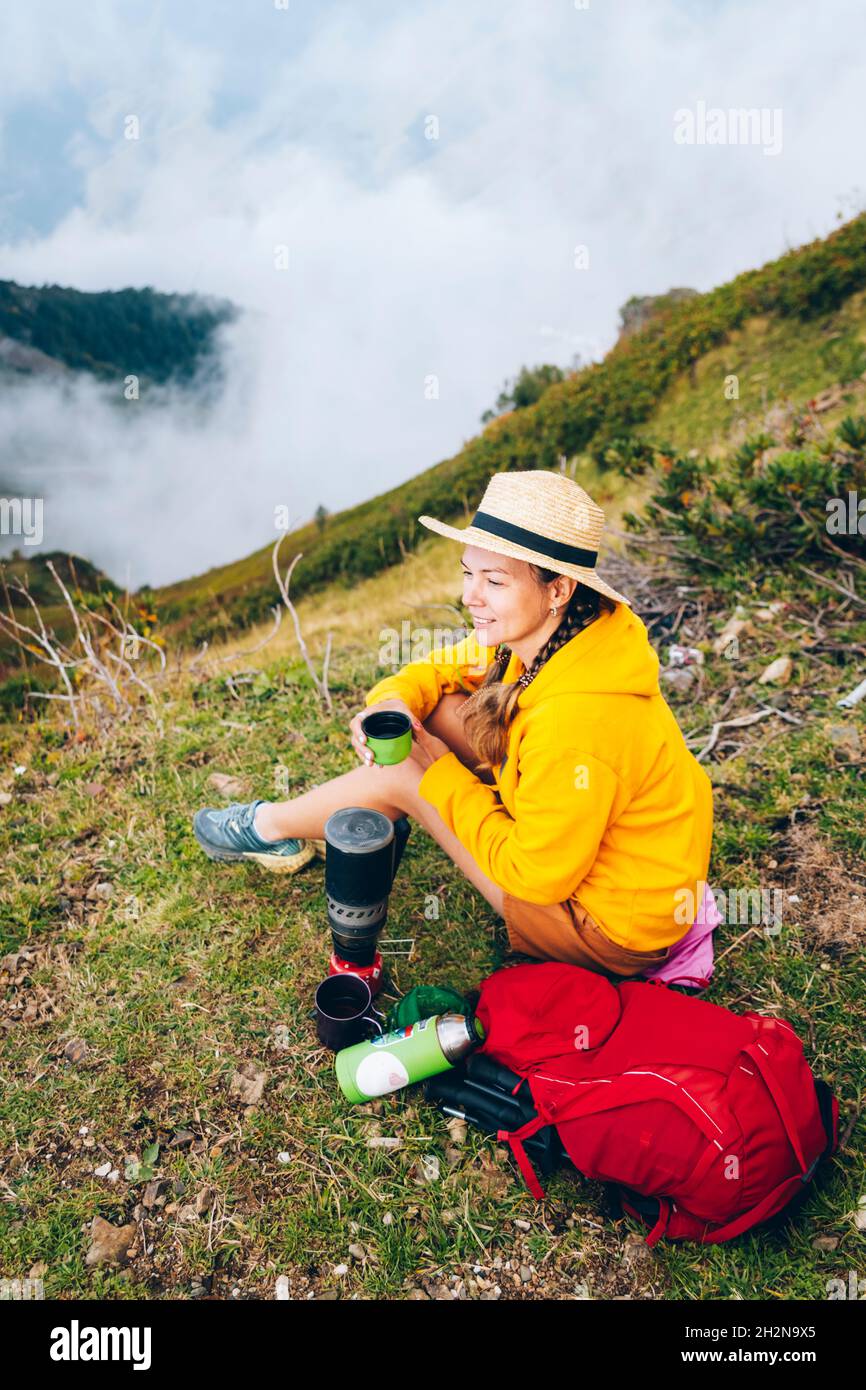 Donna sorridente che indossa cappello seduto con zaino e camping stufa montagna Foto Stock