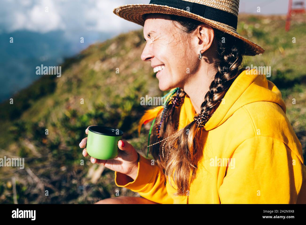 Allegra donna di metà adulta che indossa un cappello seduto in montagna durante la giornata di sole Foto Stock