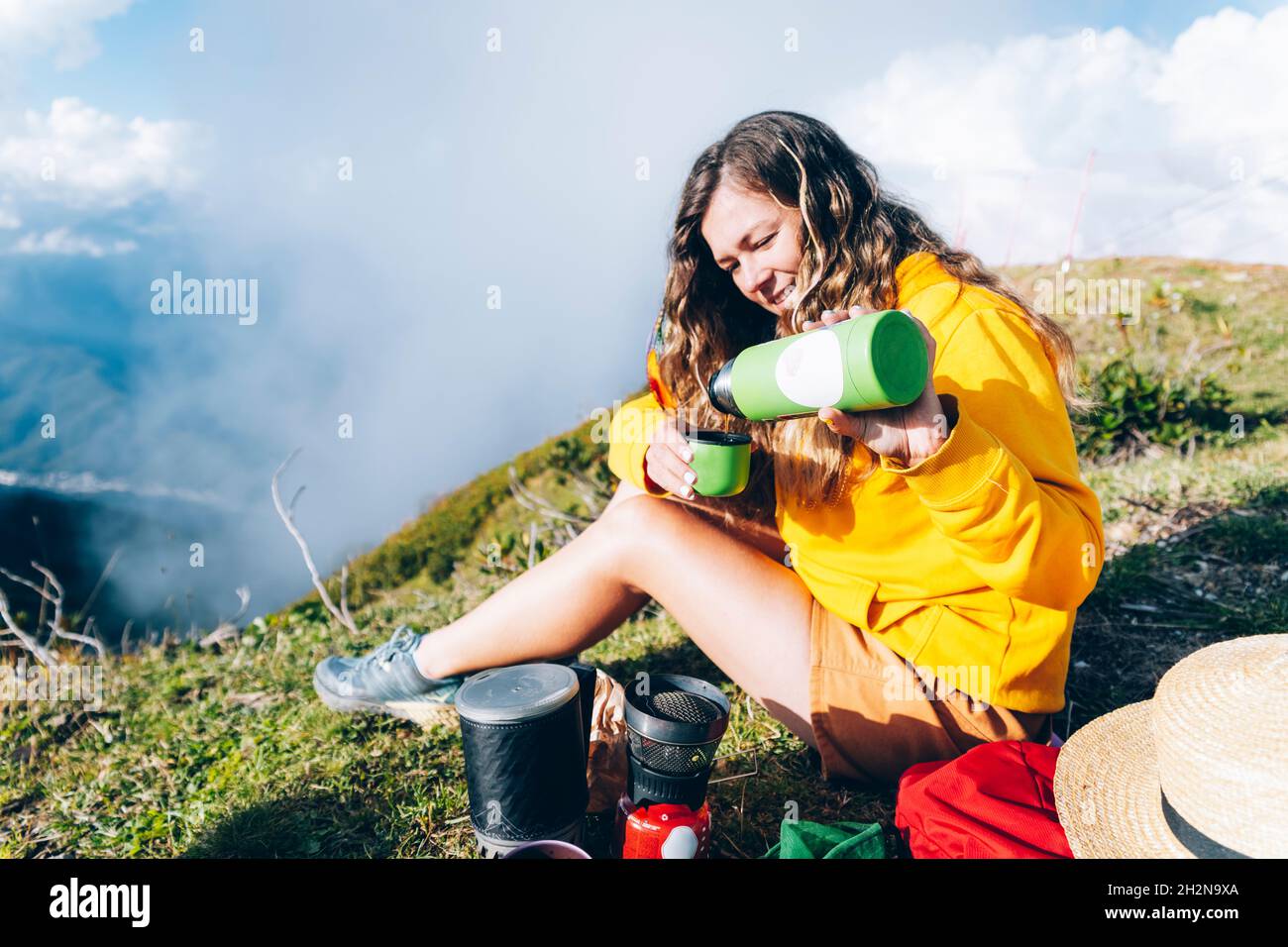 Donna che versa la bevanda dal contenitore isolato della bevanda mentre si siede sulla montagna durante la giornata di sole Foto Stock