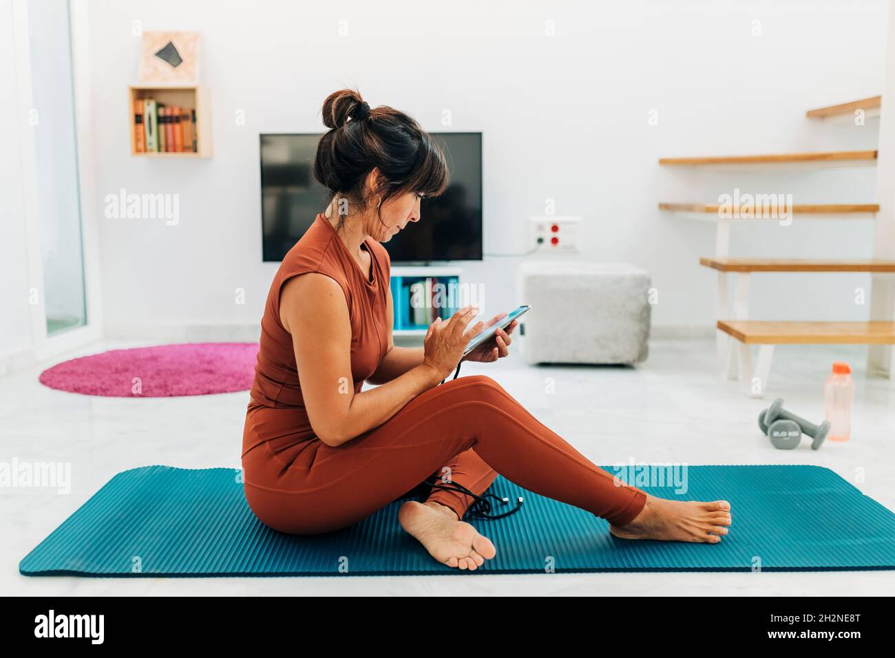 Messaggi di testo per donne mature tramite smartphone mentre si siede sul materassino per esercizi in salotto Foto Stock