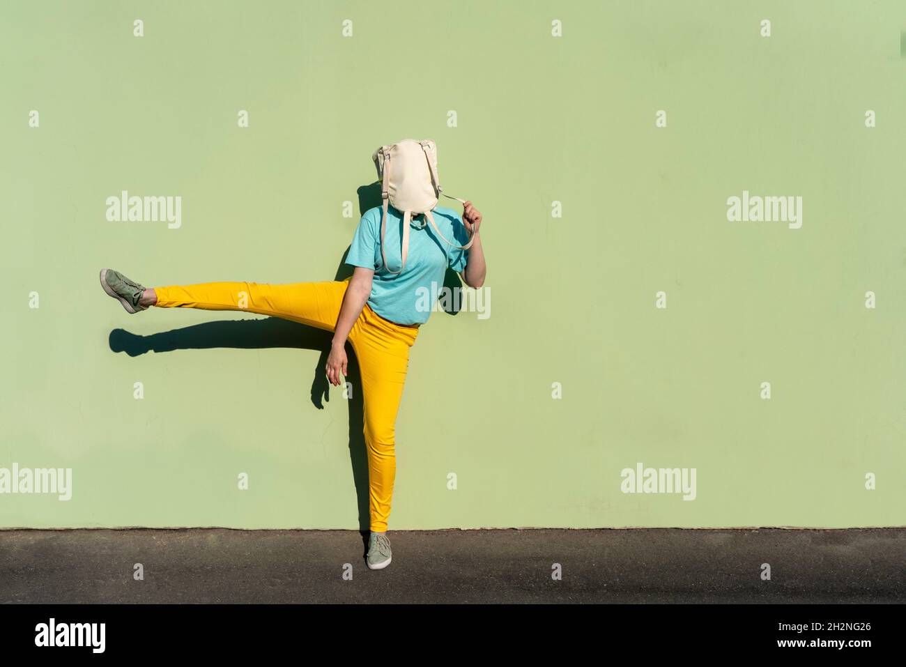 Donna di metà età adulta che ricopre il viso con zaino in piedi su una gamba accanto alla parete verde Foto Stock