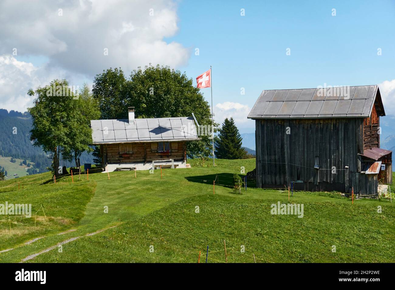 Chalet con una bandiera svizzera ondeggiante e un vecchio fienile su Un prato verde. Nello sfondo nuvole e cielo blu. Surselva Grigioni Svizzera Foto Stock