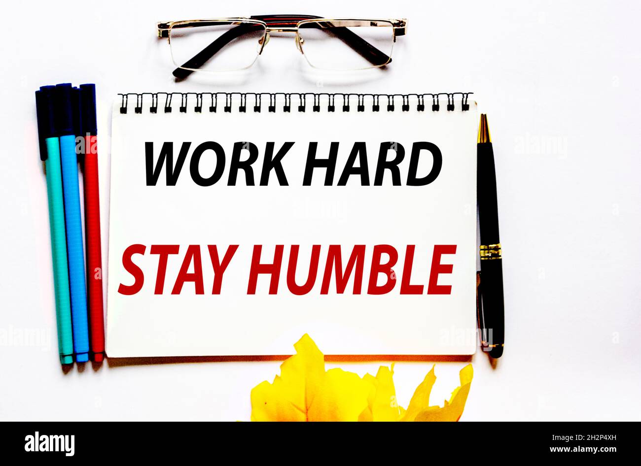 Lavorare lhard, rimanere umili, il testo è scritto su un notebook bianco, marcatori e occhiali sono accanto ad esso. Foto Stock
