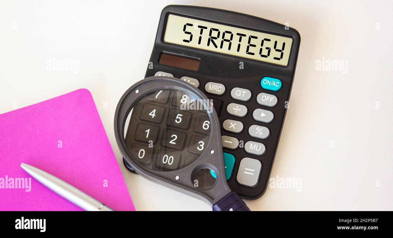 LA parola STRATEGIA su un display calcolatore, vicino ad una lente di ingrandimento e notepad su uno sfondo bianco Foto Stock